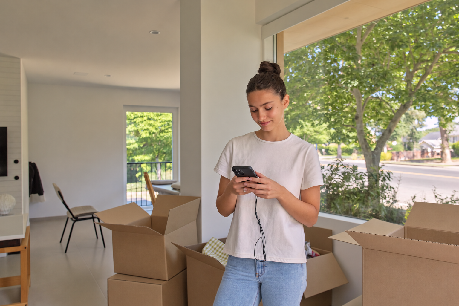Junge Frau steht in einem hellen, fast leeren Zimmer zwischen Umzugskartons und blickt lächelnd auf ihr Smartphone